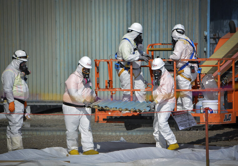 Workers move a transite siding panel from the X-326 Process Building in preparation for wrapping and removal.